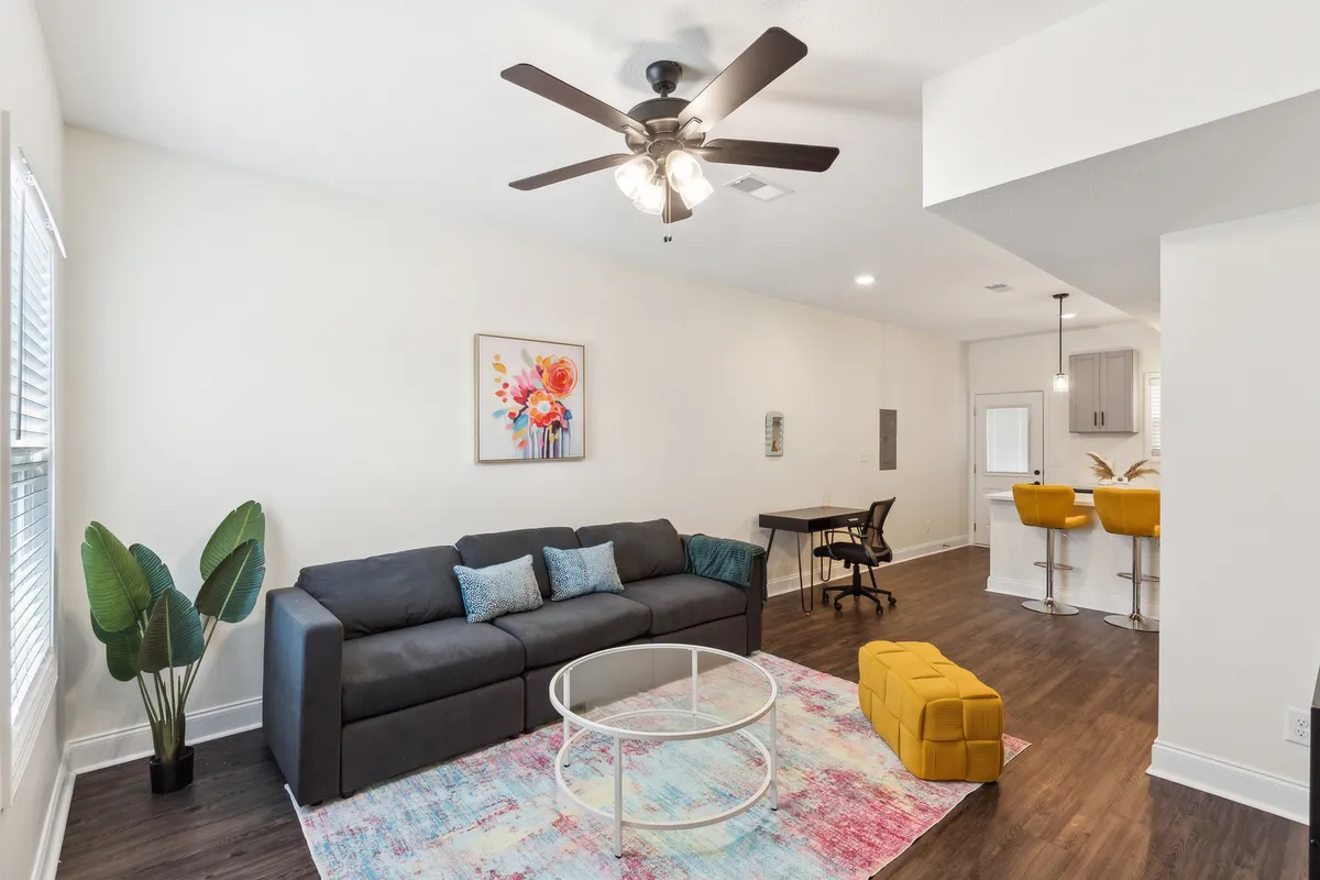 The image showcases a bright, contemporary living space featuring a plush sofa, stylish coffee table, vibrant yellow ottoman, modern artwork, and a dedicated desk area. The room exudes warmth and comfort, blending spaces for relaxation, socializing, and working.