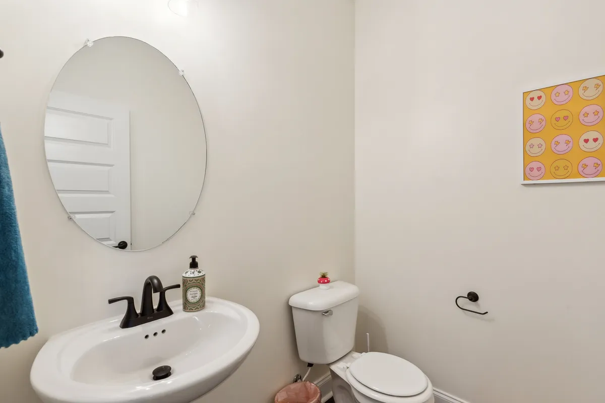 A stylish half bath featuring a modern oval mirror, a sleek sink, a pop of color from playful artwork, and a neatly hung towel. The space feels fresh and welcoming—perfect for guests.