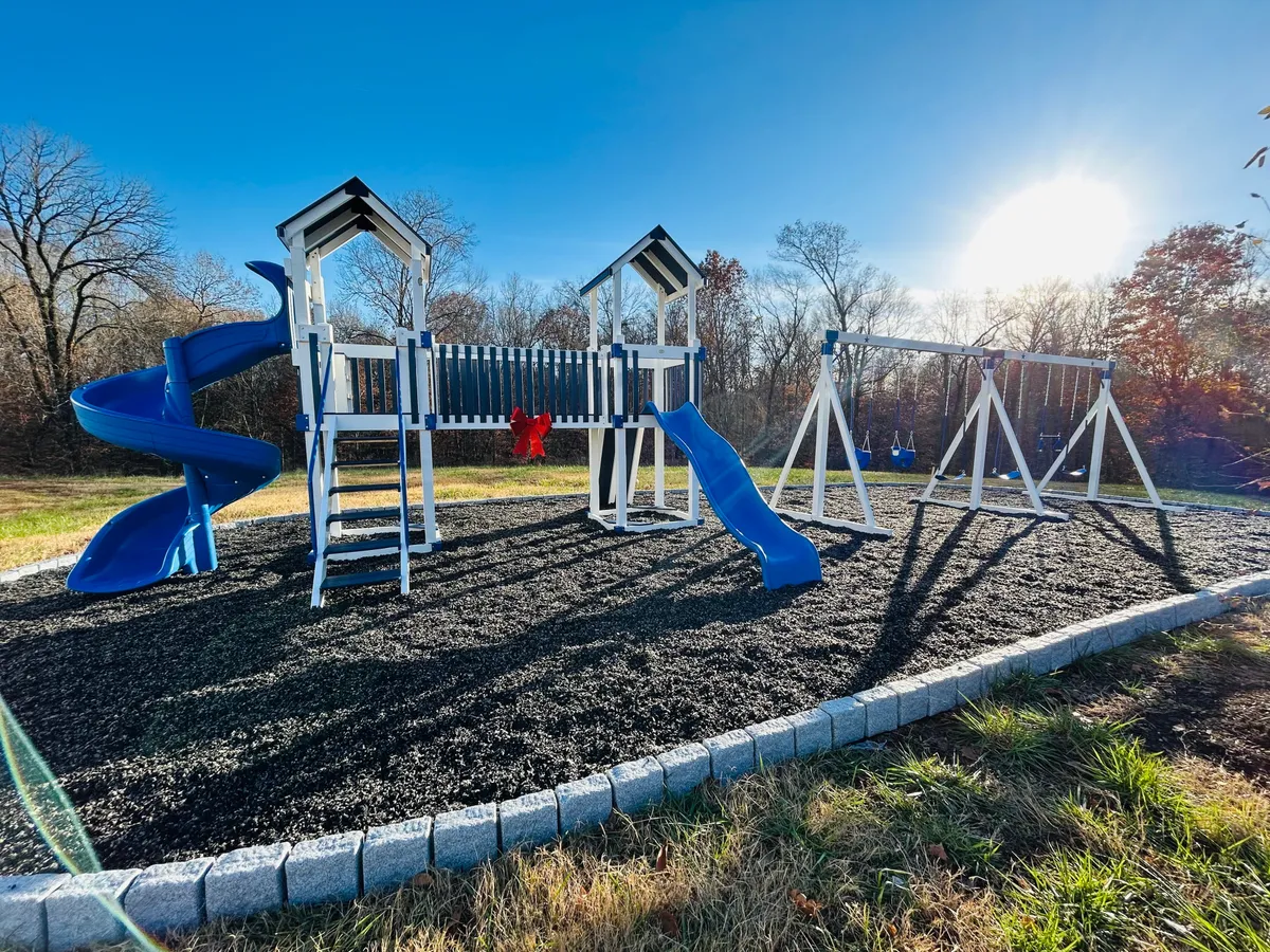 A vibrant outdoor playground featuring a blue slide, swing set, and cheerful red decorations, set in an inviting green space. Children can be seen playing and families are gathered nearby, enjoying picnics and quality time together in the fresh air.