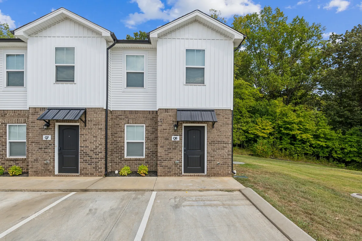 An inviting exterior view of a two-story townhouse with neatly landscaped areas in front. The image captures the entrances, front doors, and a communal outdoor space where residents can be seen socializing and enjoying the neighborhood. Vehicles are parked conveniently close, highlighting ease of access.