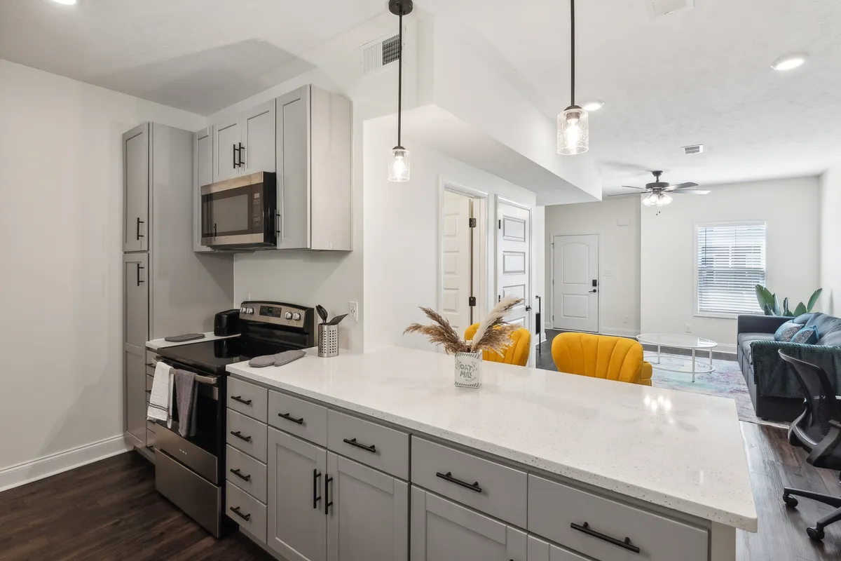 A bright and welcoming kitchen is shown with a central island, yellow accent chair, and modern appliances. The open-plan design encourages socializing, cooking, and entertaining, seamlessly connecting with adjacent zones for a communal feel.