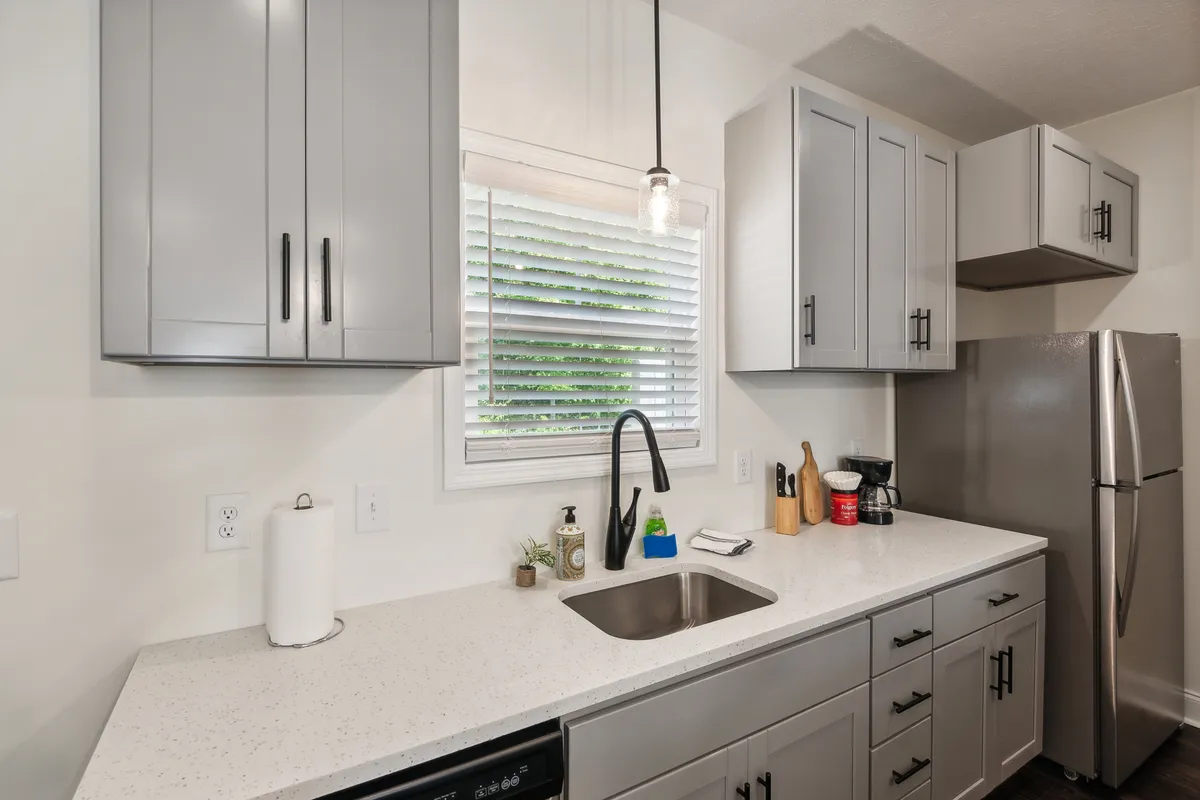 The image displays a contemporary kitchen sink area with sleek countertops, cabinets, and a window that brings in natural light. The space is functional yet inviting, supporting activities like cooking, family gatherings, and morning routines.