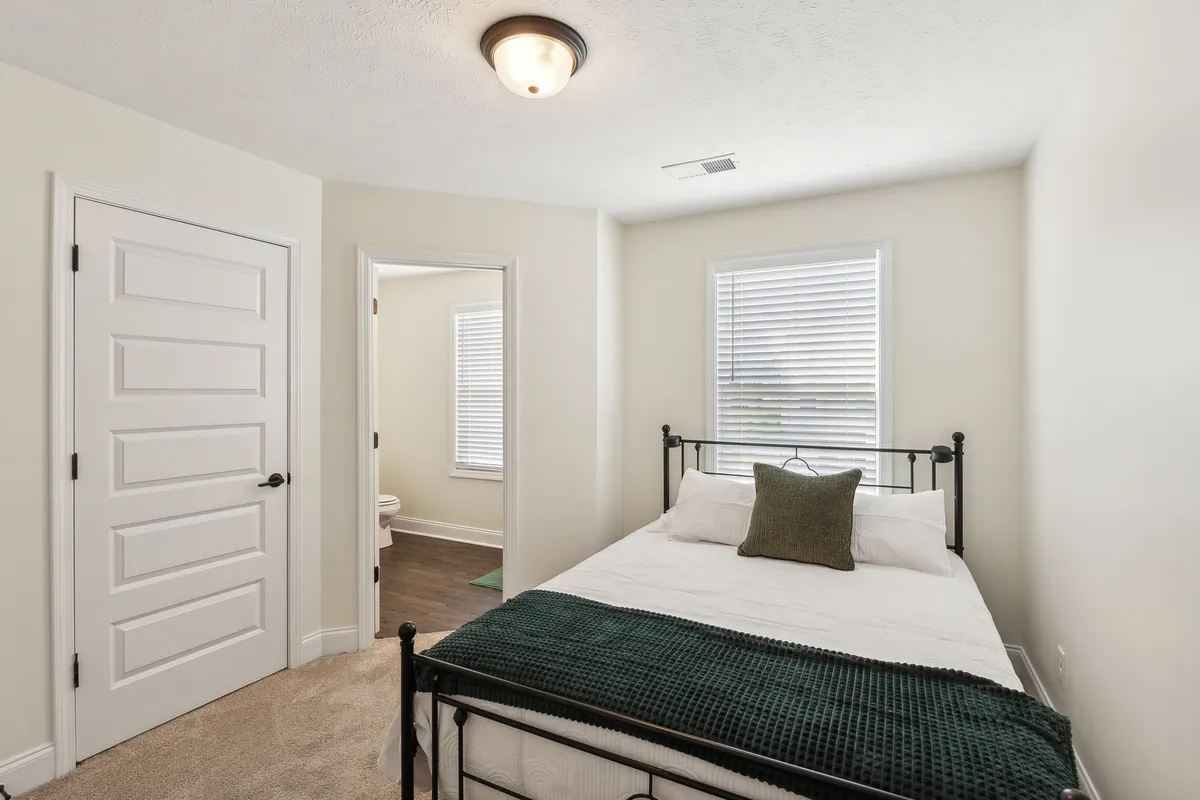 A bedroom with a comfortable bed draped in a green throw, windows with blinds letting in soft natural light, and a doorway leading to an adjoining bathroom. The room offers both tranquility and a practical layout for privacy.