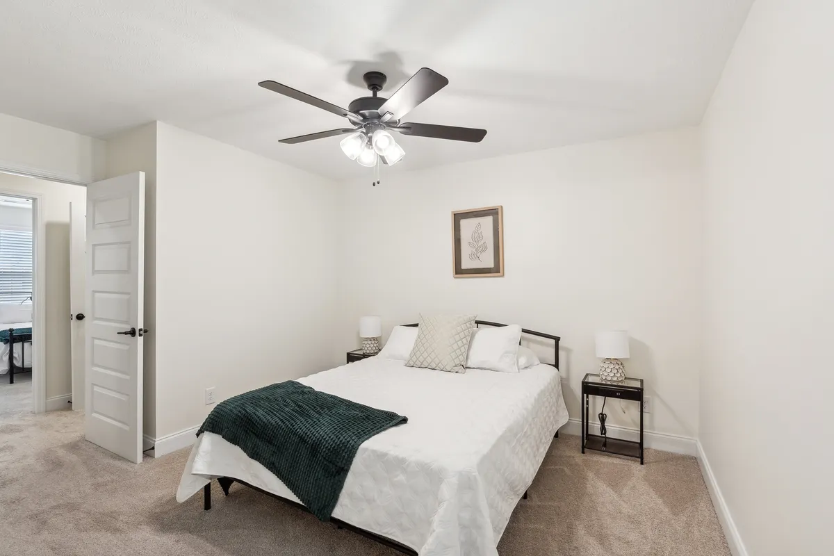 A serene bedroom featuring a bed with a decorative pillow, matching nightstands with warm-lit lamps on either side, a ceiling fan overhead, and tasteful wall art. The overall ambience is restful and inviting, suggesting a comfortable retreat after time spent out exploring.